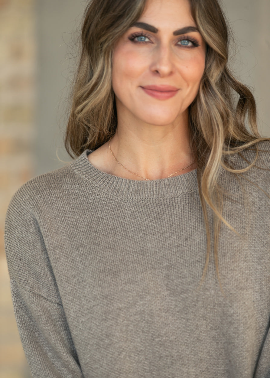 A woman with blonde-highlighted hair wearing a cozy, heathered taupe-gray knit crewneck sweater, shown in a close-up portrait.