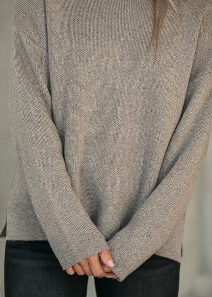 A close-up of a person wearing an oversized, long-sleeve knit sweater in a heathered taupe-gray color, paired with dark jeans.