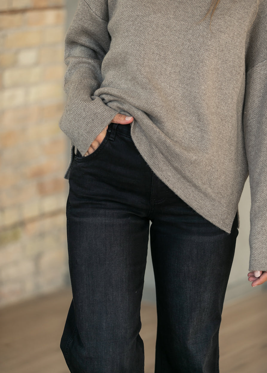 A person wearing an oversized taupe knit sweater and dark charcoal jeans, standing in front of a softly blurred brick wall.