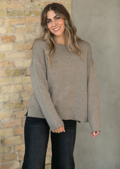 A woman wearing a light taupe knit sweater and dark jeans, standing in front of a split brick and concrete wall.