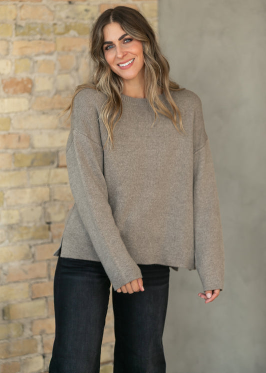 A woman wearing a light taupe knit sweater and dark jeans, standing in front of a split brick and concrete wall.
