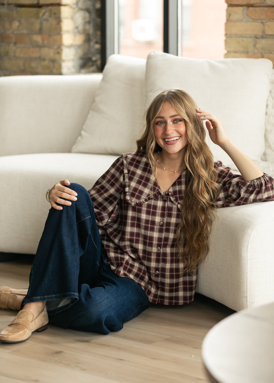 A woman with long wavy light brown hair sits on a light wood floor in front of a white sofa, wearing a brown and burgundy plaid button-up shirt, dark blue jeans, and tan loafers.