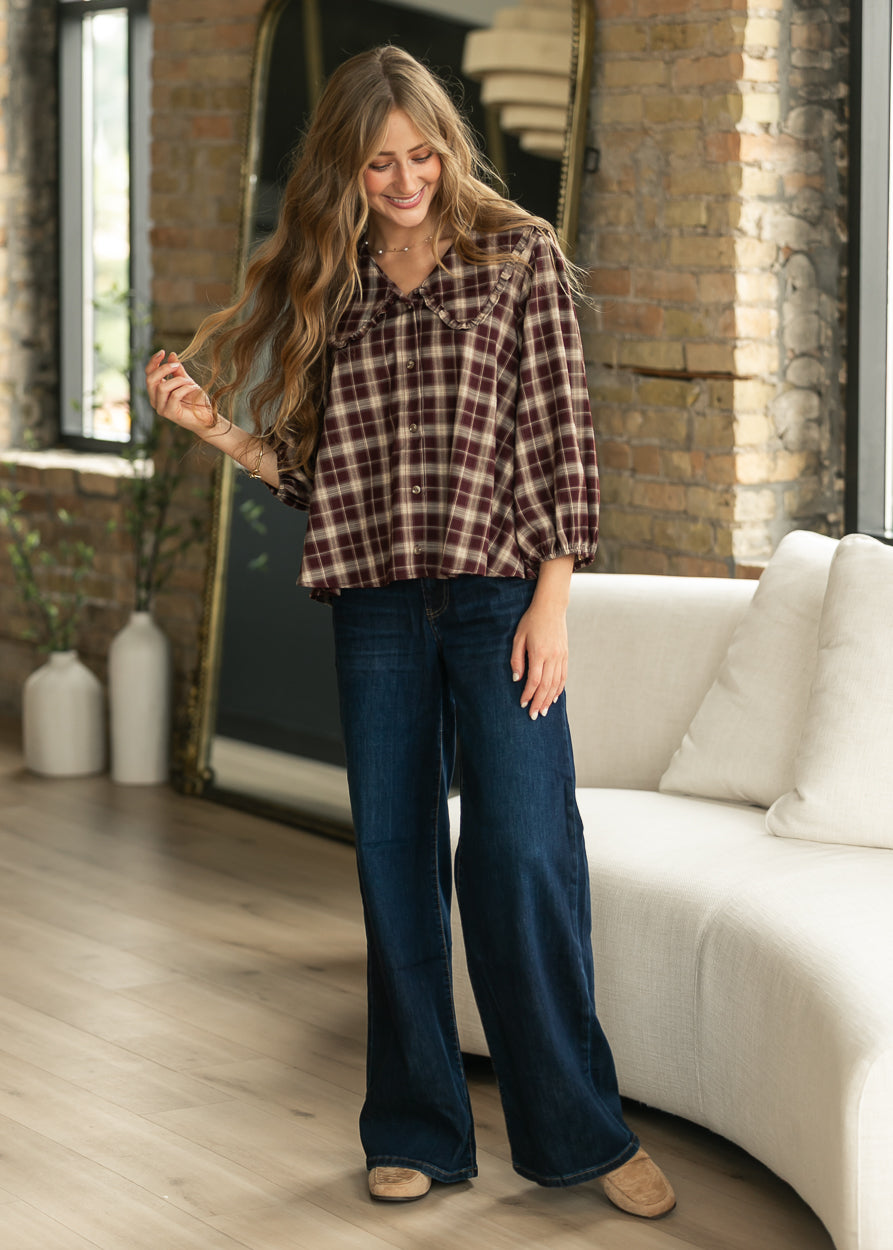 A woman stands in a bright living room wearing a brown and red plaid button-up shirt with dark blue wide-leg jeans and tan shoes.