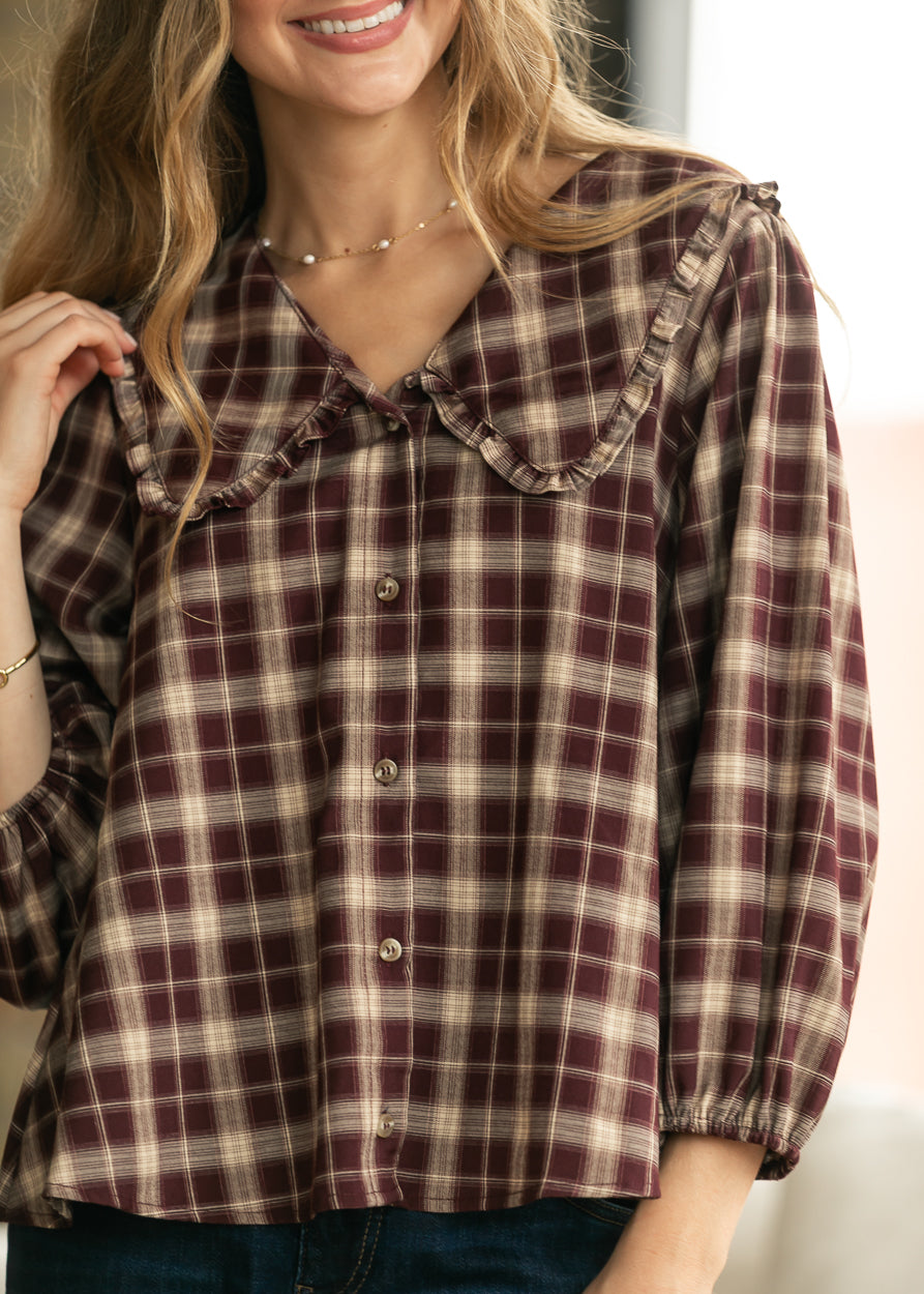 Close-up of burgundy plaid blouse showing ruffle-trim V-neckline, buttons, and gathered sleeves.