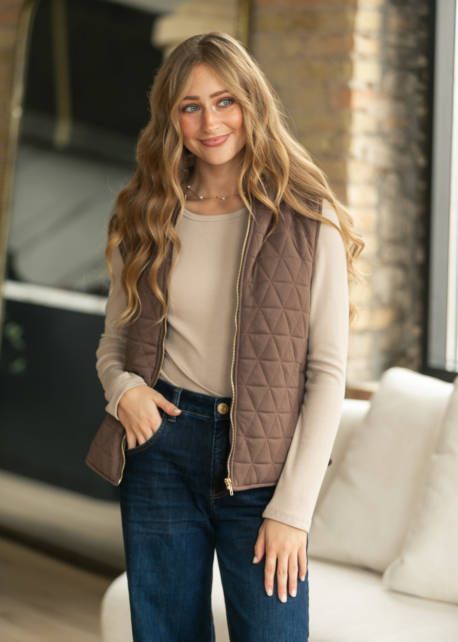 A woman wearing a taupe-brown diamond-quilted zip-front vest over a beige long-sleeve top and dark blue jeans, standing in a modern indoor setting with a blurred brick wall and white sofa in the background.
