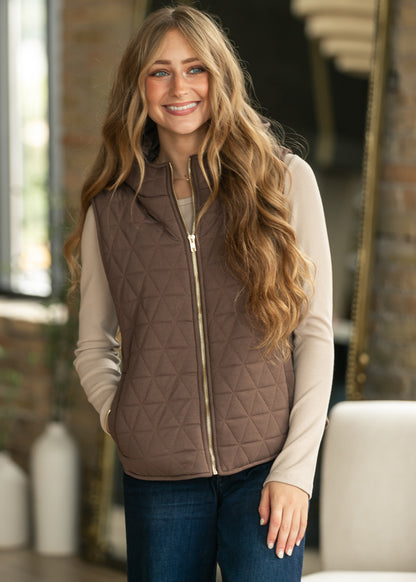 A smiling woman with long wavy light brown hair models a brown diamond-quilted zip-up vest over a beige long-sleeve shirt and blue jeans in an indoor setting with a blurred brick wall background.