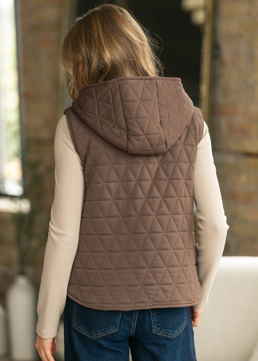 A woman viewed from behind wearing a sleeveless, quilted brown hooded vest over a cream long-sleeve shirt and blue jeans, standing in an indoor setting with a brick wall background.