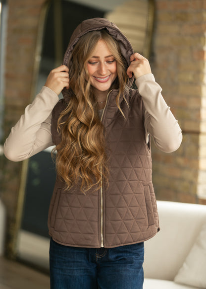 A woman wearing a brown diamond-quilted zip-front hooded vest over a light beige long-sleeve shirt and blue jeans, standing in a softly lit indoor setting with a brick wall and white sofa in the background.