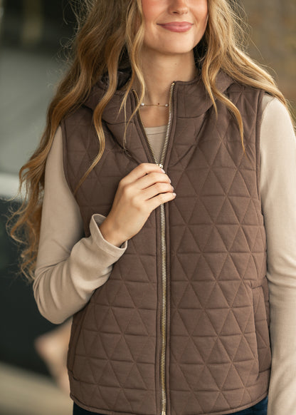 A woman wearing a brown diamond-quilted zip-up puffer vest over a light beige long-sleeve top, shown from the chest up in a softly blurred outdoor setting.
