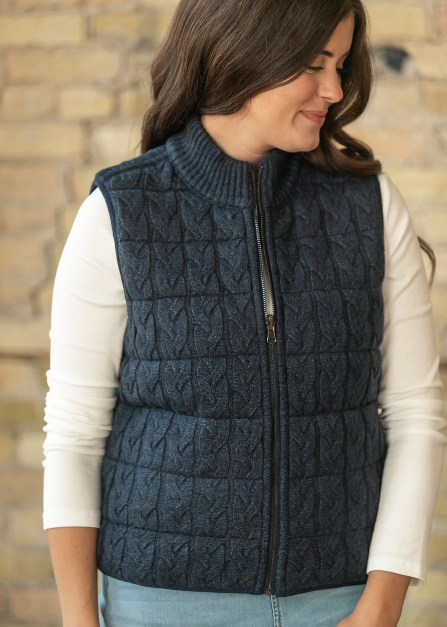 Navy reversible cable-knit vest with ribbed mock neck and full zip, layered over a cream long sleeve top.