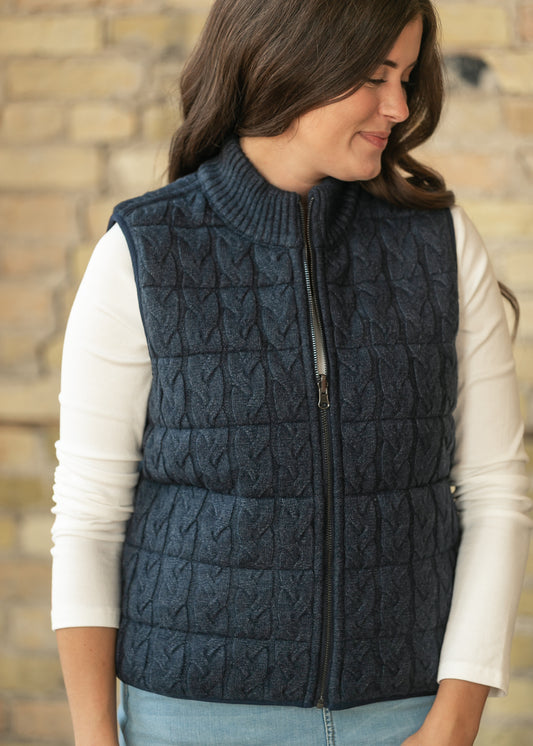 Navy reversible cable-knit vest with ribbed mock neck and full zip, layered over a cream long sleeve top.