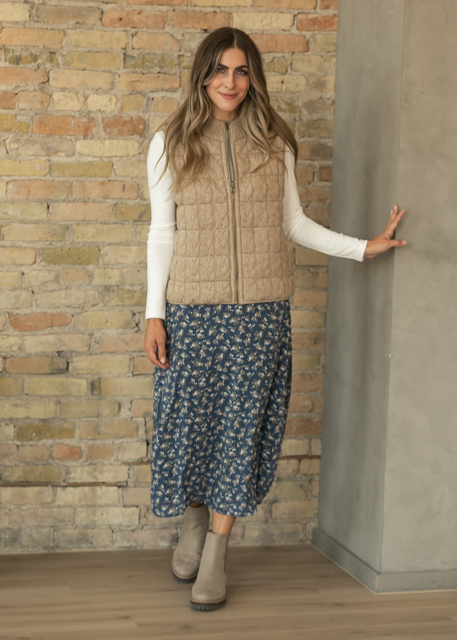 Taupe cable-knit vest styled with a cream long sleeve top, navy floral midi skirt, and neutral ankle boots.