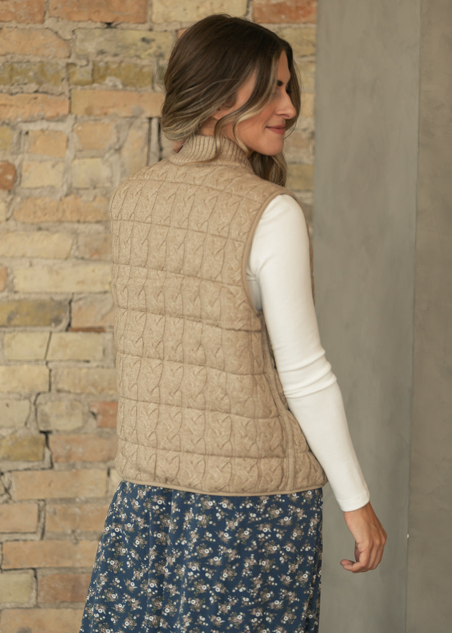 Back of taupe reversible cable-knit vest layered over a cream long sleeve top and navy floral skirt.