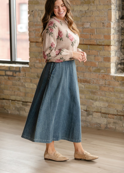 Sand pink rose sweater styled with an A-line denim midi skirt and tan suede flats beside a window.