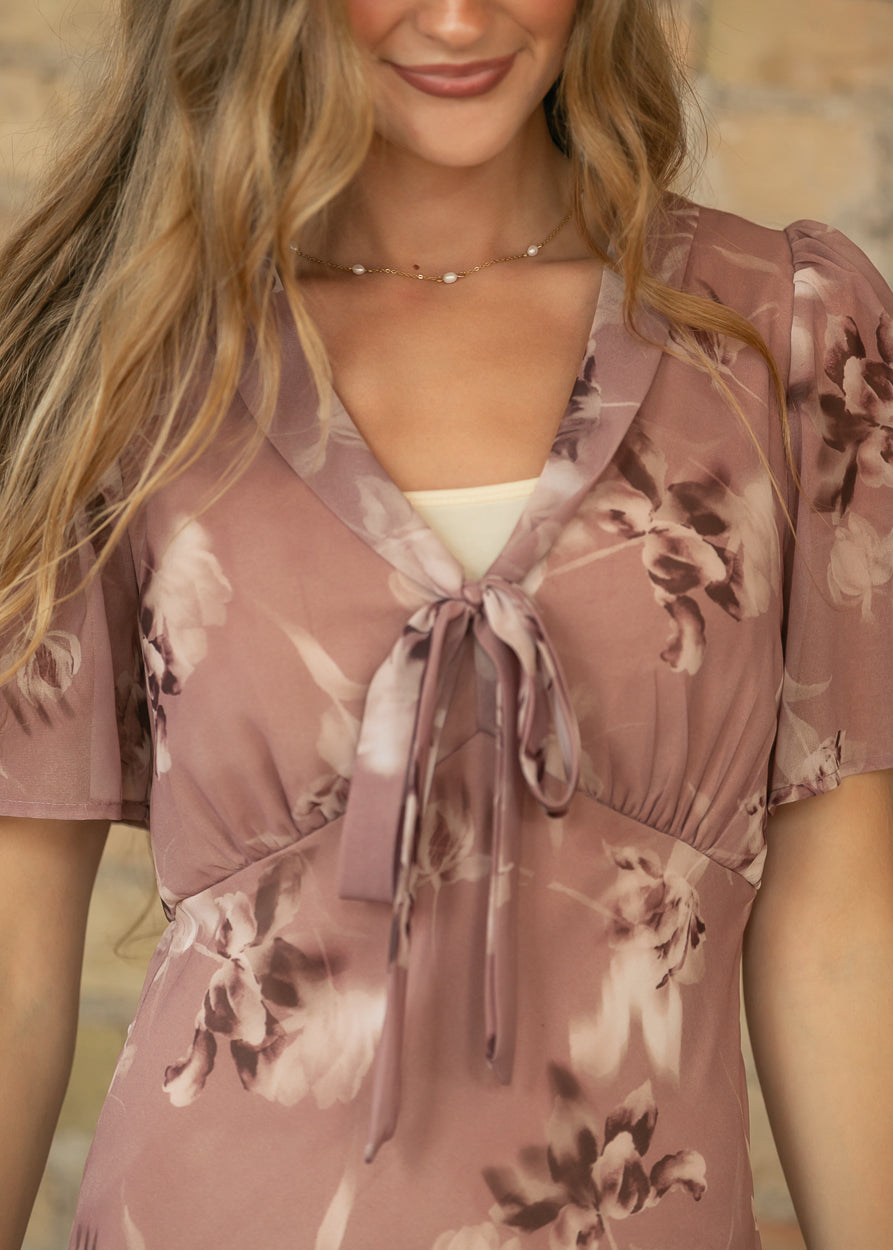 Close-up of mauve floral maxi dress showing V-neckline, front tie detail, gathered bodice, and short sheer sleeves.