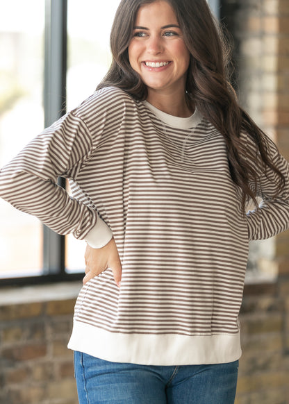 A woman wearing a brown and cream horizontally striped long-sleeve crewneck sweatshirt with solid cream ribbed trim, paired with blue jeans, standing in front of a brick wall and window.