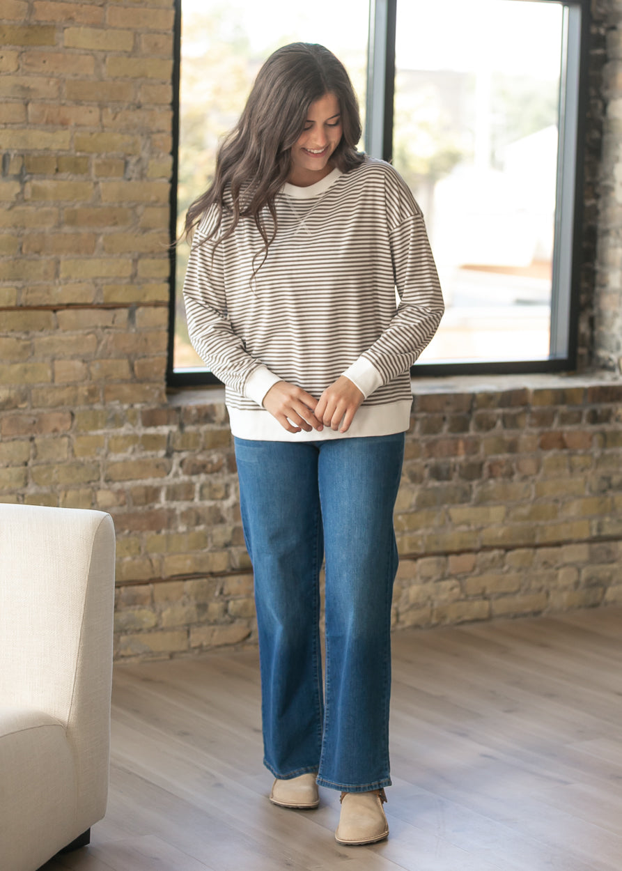A woman wearing a beige and white striped long-sleeve top with blue jeans and beige shoes, standing in a room with a brick wall and large window.