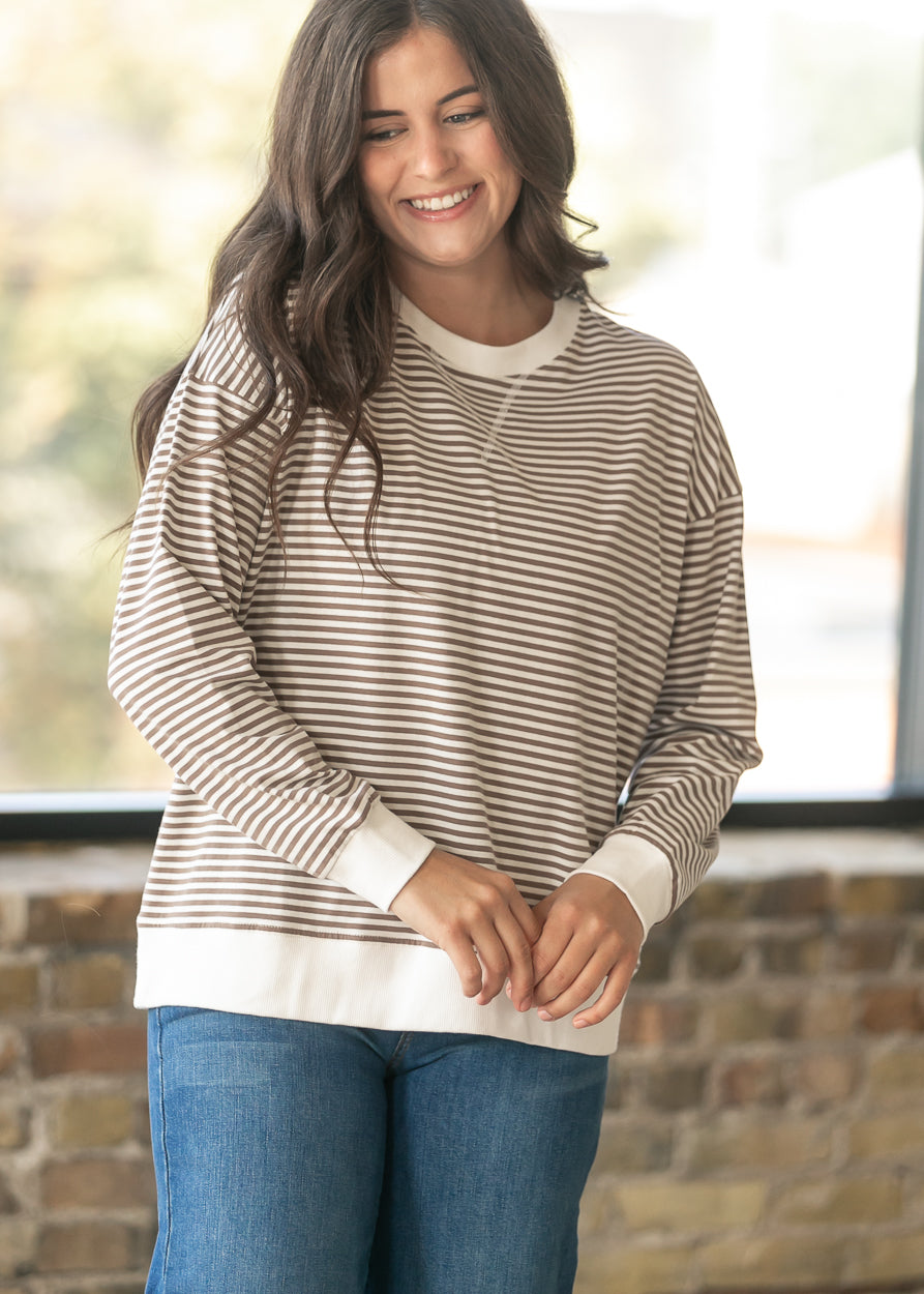 A woman wearing a beige and white horizontally striped long-sleeve crewneck sweatshirt with solid white ribbed trim, paired with blue jeans, standing in front of a brick wall and window.