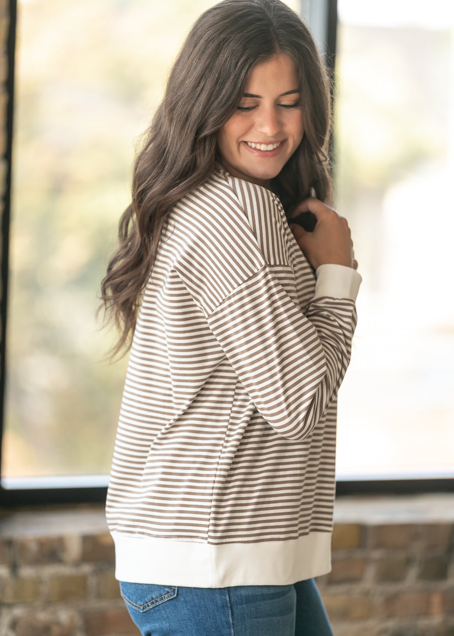 A woman with long brown hair is shown from the side wearing a brown and cream horizontally striped long-sleeve sweatshirt with solid cream ribbed cuffs and hem, paired with blue jeans.