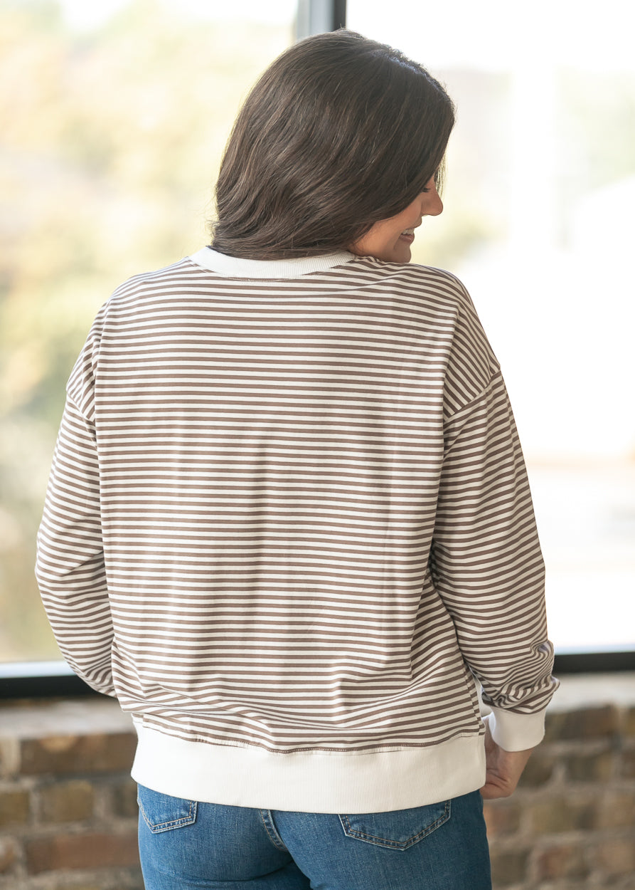 A woman viewed from behind wearing a beige and white horizontally striped long-sleeve sweatshirt with solid white ribbed cuffs and hem, paired with blue jeans.
