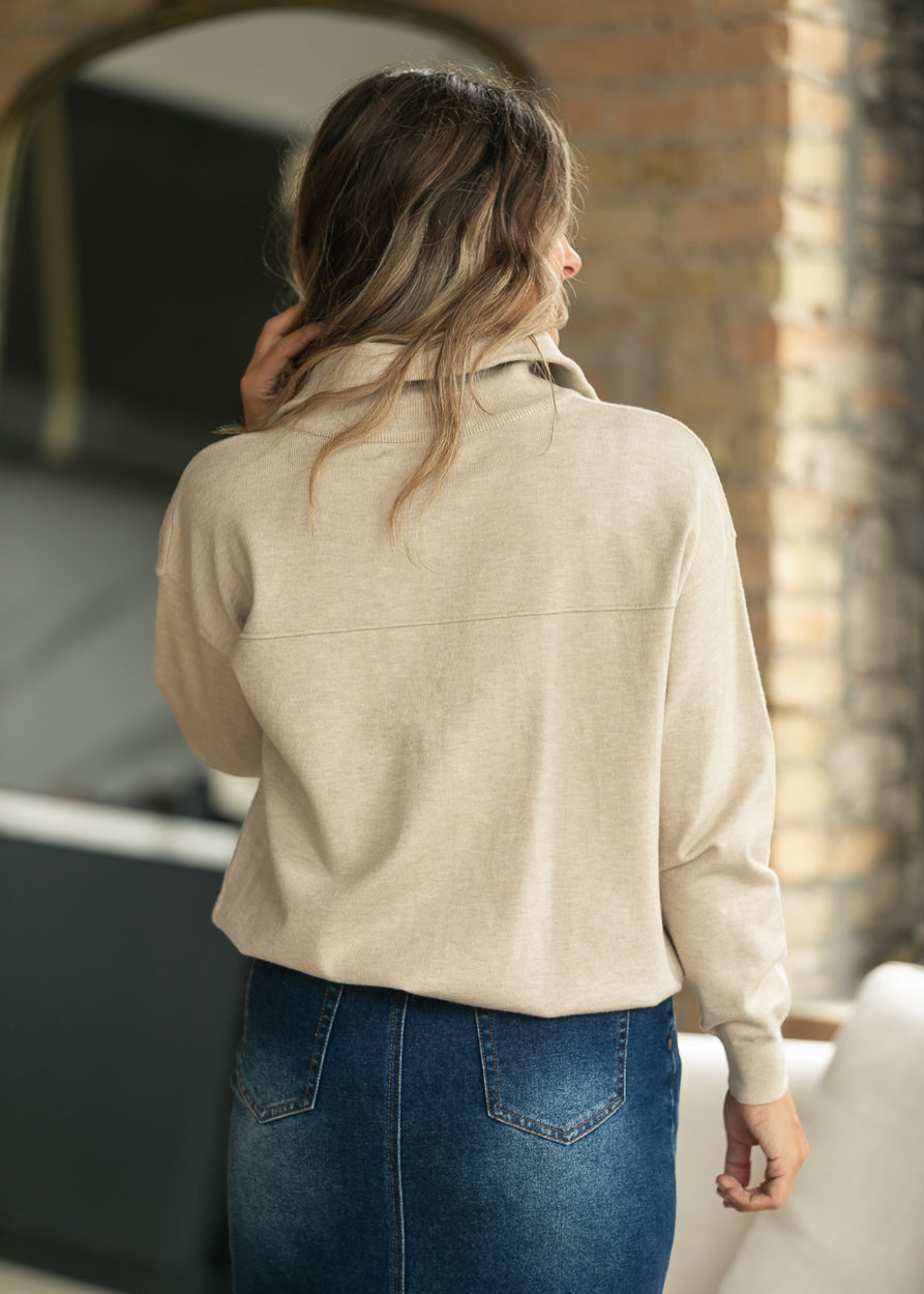 A woman viewed from behind wearing a light beige knit long-sleeve top and a blue denim skirt in an indoor setting with a brick wall background.