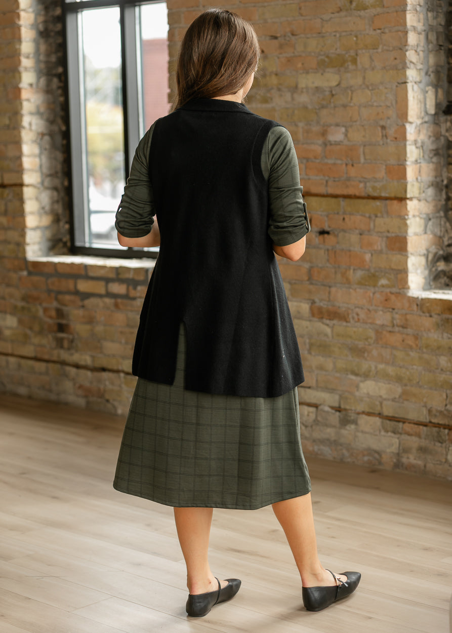 Back of olive windowpane midi dress layered with a long black sleeveless cardigan and black flats.