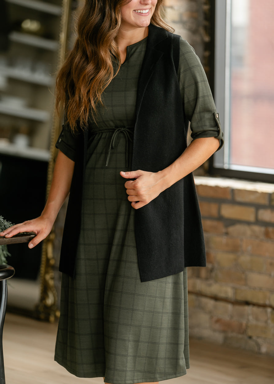 Olive windowpane dress with button-tab sleeves and a tie waist, worn under an open black cardigan.