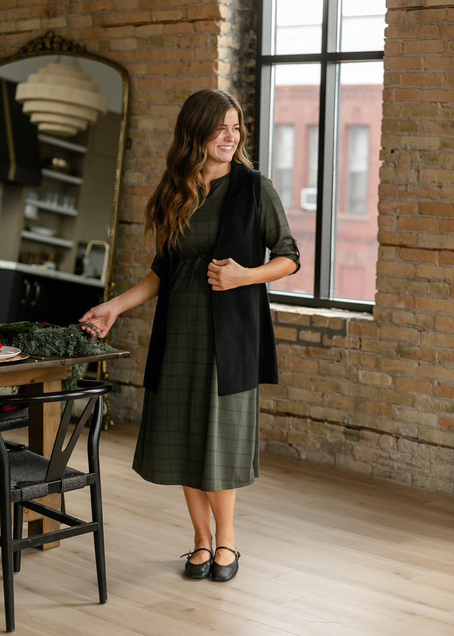 Olive windowpane midi dress styled with a black sleeveless cardigan and Mary Jane flats beside a decorated table.