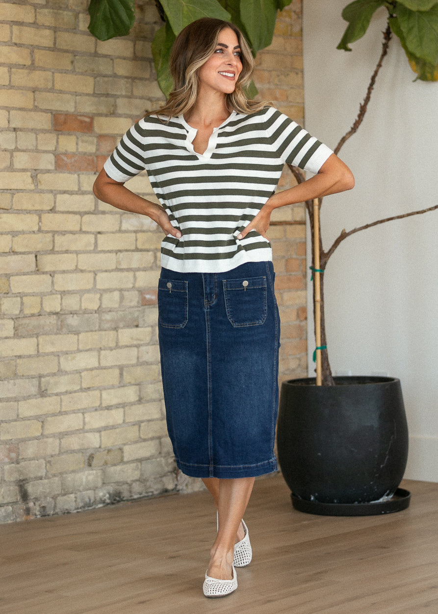 A woman modeling a short-sleeve striped knit top and a dark blue denim midi skirt, standing in a room with a brick wall, wood floor, and a potted plant.