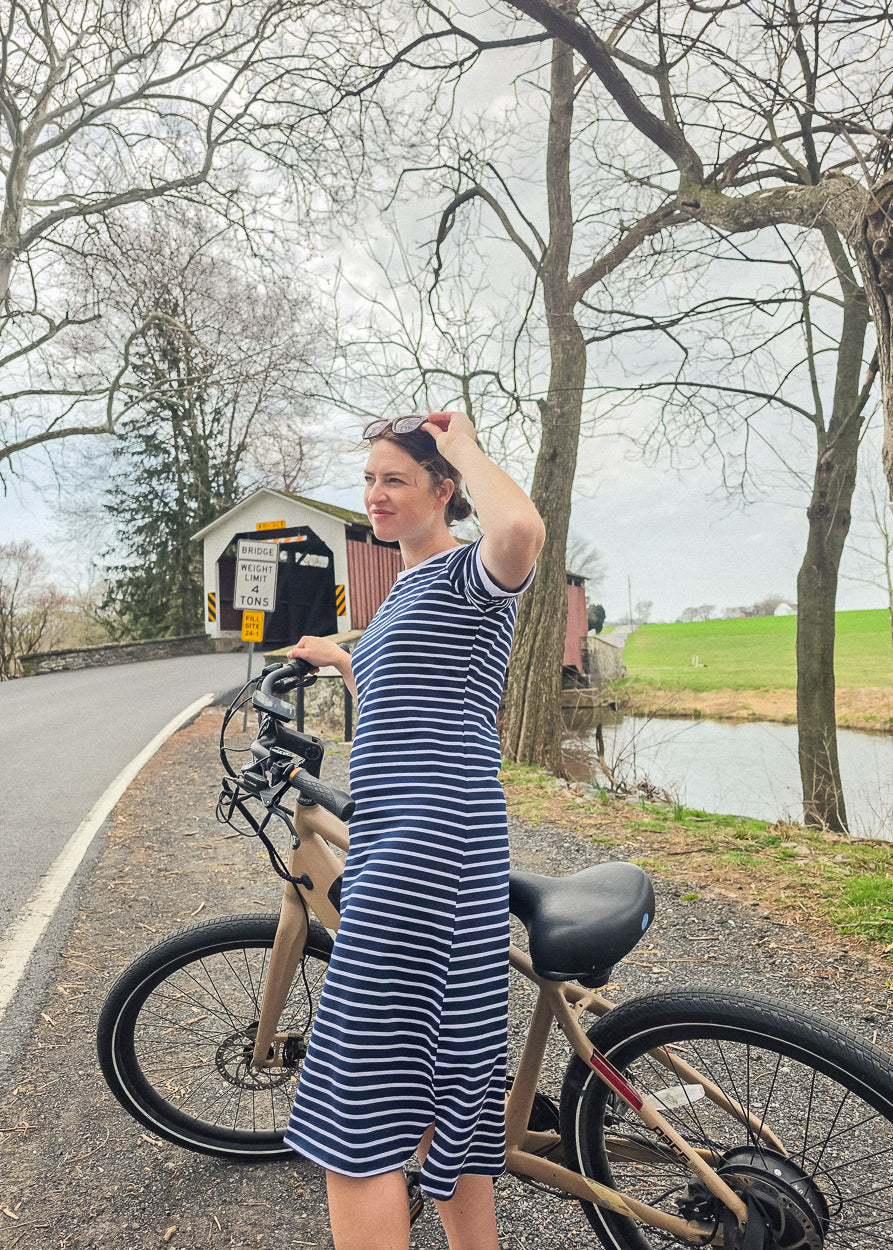 Woman in a striped dress standing next to a bicycle on a road with trees and a covered bridge in the background.