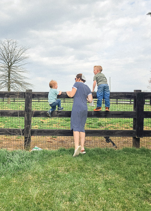 Woman in navy and white striped midi T-shirt dress with short raglan sleeves with two children by a wooden fence in a rural setting
