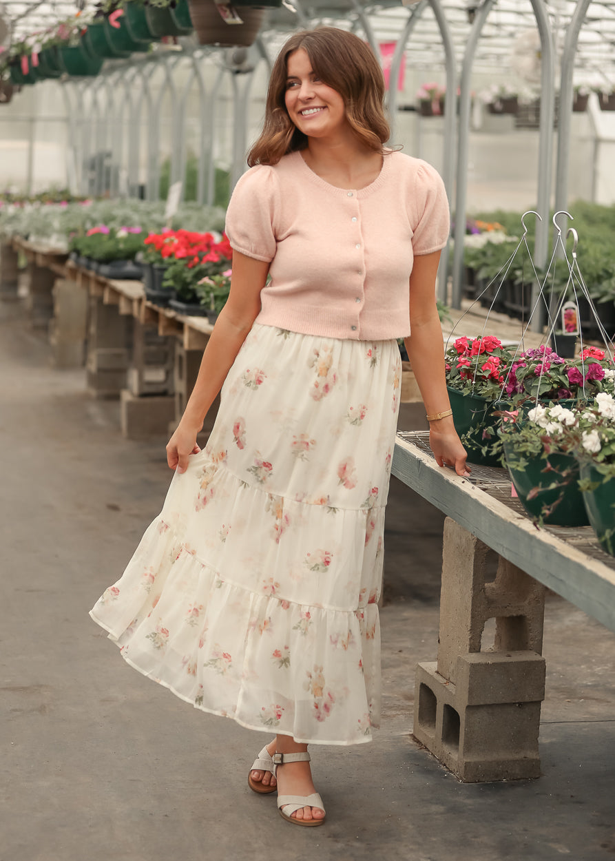 Soft pink cropped button front cardigan with puff sleeves, styled with a floral cream tiered maxi skirt and sandals.