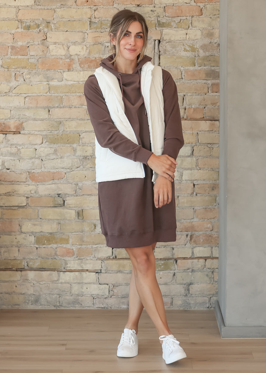 A woman modeling a brown hooded sweatshirt dress with a white faux-fur vest layered over it, standing in front of a rustic brick wall.