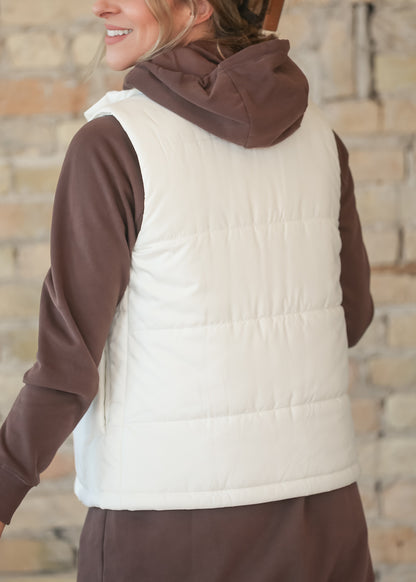 A woman wearing a cream quilted puffer vest over a brown hooded sweatshirt and brown pants, photographed from the back against a blurred light brick wall.