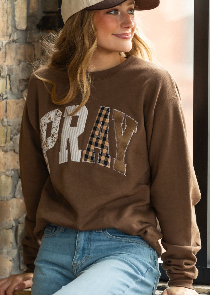 Brown PRAY graphic sweatshirt styled with light wash jeans and a neutral cap