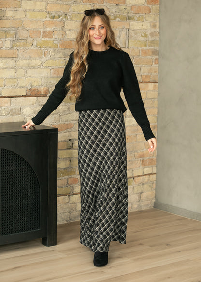 A woman stands indoors wearing a black long-sleeve knit top and a long black-and-white diamond-patterned skirt, posed in front of a brick wall and wood floor.