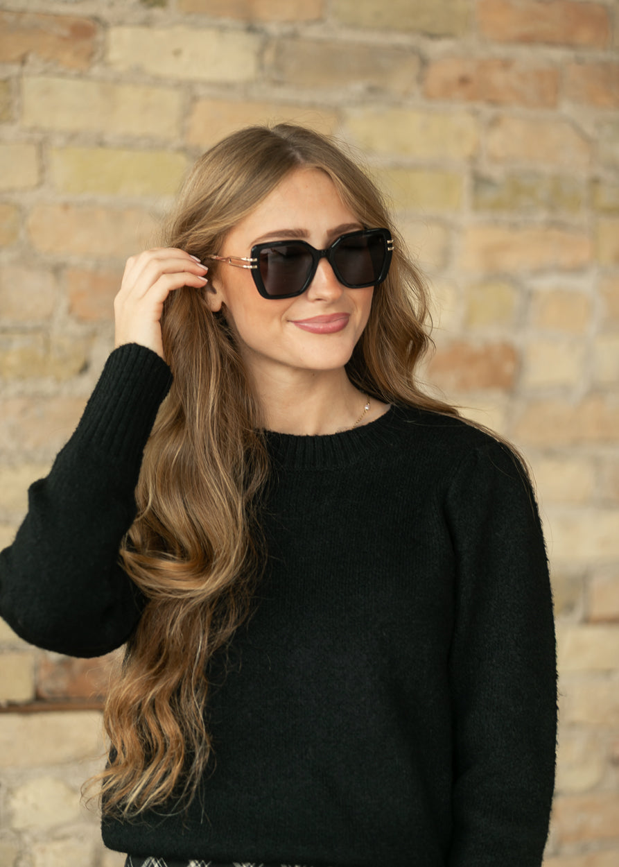 A woman with long wavy light brown hair wearing black sunglasses and a black knit sweater, posing in front of a rustic brick wall.