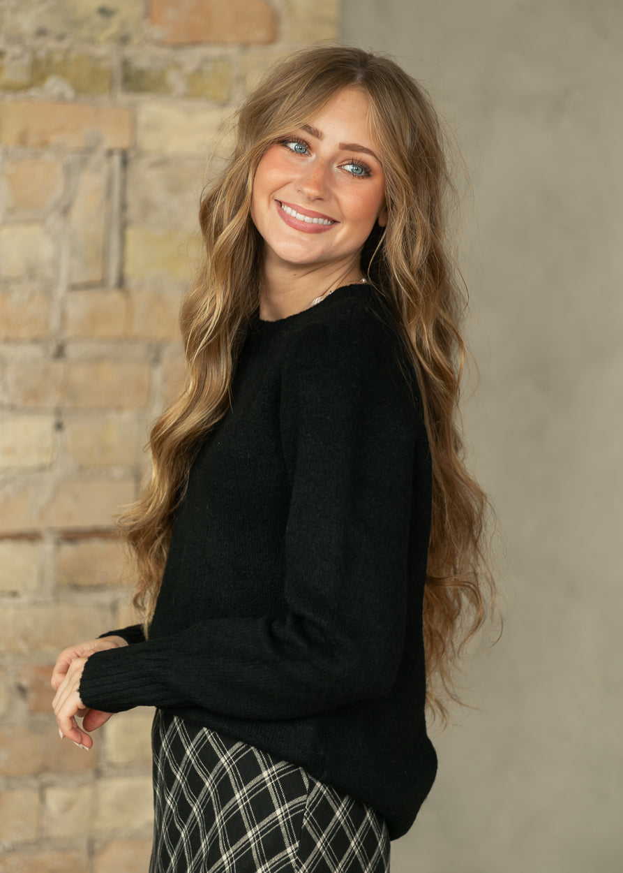 A smiling woman with long wavy light brown hair wearing a black knit crewneck sweater and a black-and-white plaid skirt, standing against a rustic brick and concrete wall.