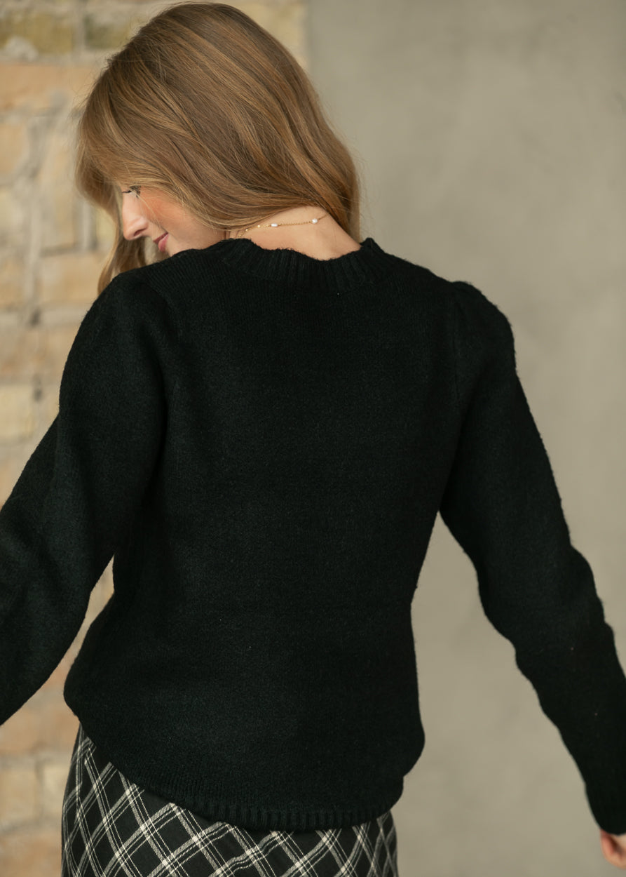 A woman wearing a black knit sweater, shown from the back, with her head turned to the side. She is wearing a black and white plaid skirt, and the background features a textured gray wall and a rustic brick section.