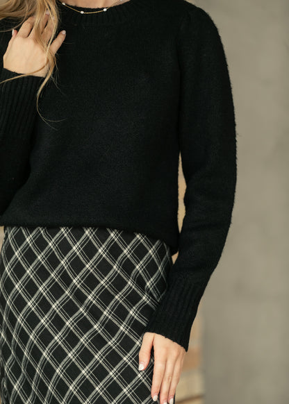 A close-up of a person wearing a black knit sweater and a black-and-white plaid skirt, with one hand touching the sweater and the other resting near the skirt.