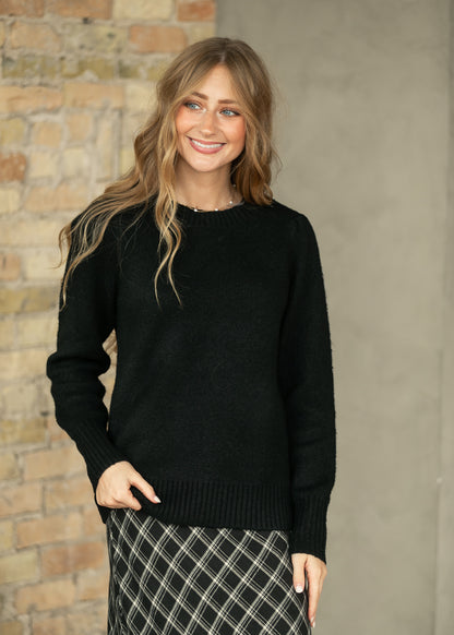 A woman wearing a black knit crewneck sweater and a black-and-white plaid skirt, standing against a split brick and concrete wall.