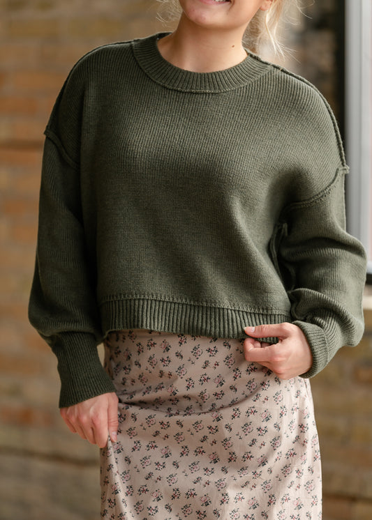 Close-up of olive green sweater showing exposed shoulder seams, ribbed neckline, and relaxed knit texture.