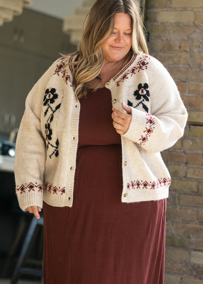 Cream geometric floral button-up cardigan with black floral motifs, burgundy knit accents, and ribbed trim, styled over a mulberry midi dress.
