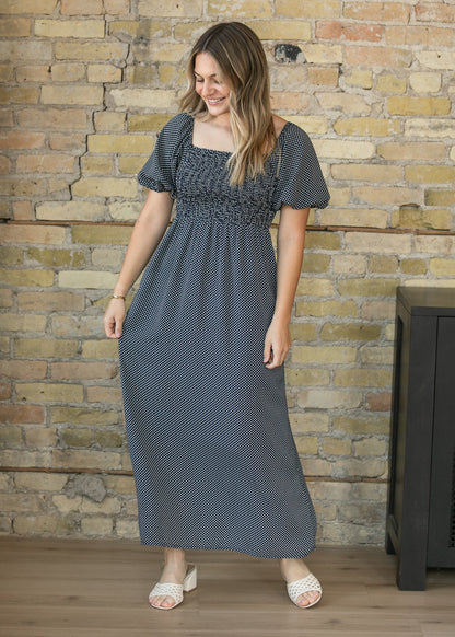 A woman wearing a dark gray and white micro-patterned maxi dress with a smocked bodice and short puff sleeves, standing in front of a rustic brick wall.