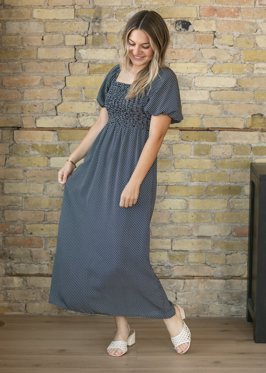 A woman modeling a dark gray and white micro-dot maxi dress with a smocked bodice, standing in front of a rustic brick wall.