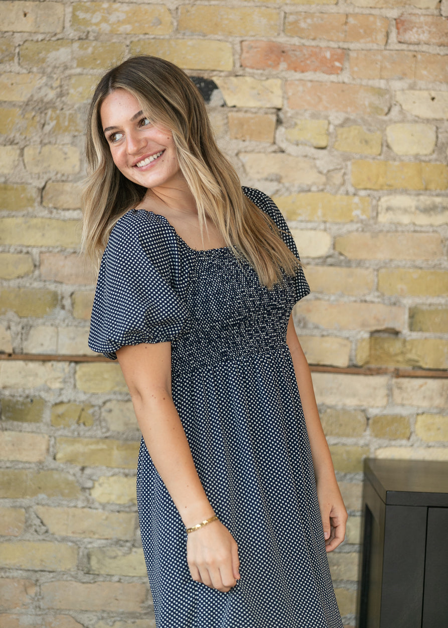 A woman with blonde-highlighted hair wearing a navy and white micro-dot dress, standing in front of a rustic yellow brick wall.