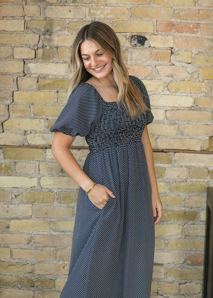 A woman wearing a navy blue and white micro-dot dress with puff sleeves and a smocked bodice, standing against a yellow brick wall.