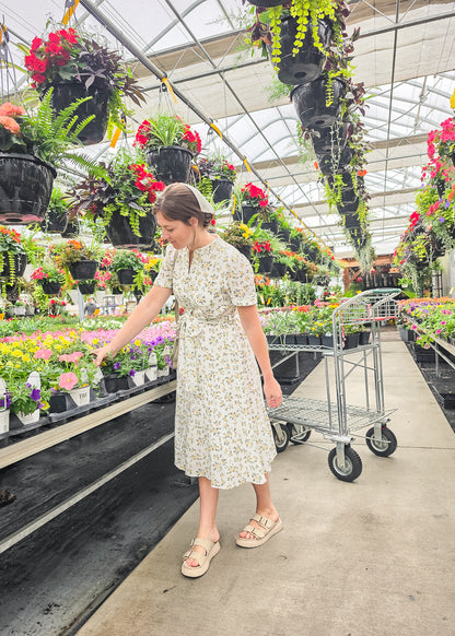 Woman in a floral dress shopping in a greenhouse filled with plants and flowers.