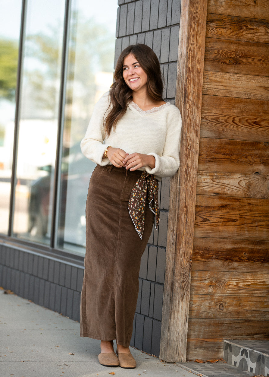 Brown corduroy maxi skirt styled with a cream sweater, floral scarf, and tan flats outdoors.
