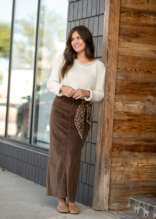 Brown corduroy maxi skirt styled with a cream sweater, floral scarf, and tan flats outdoors.